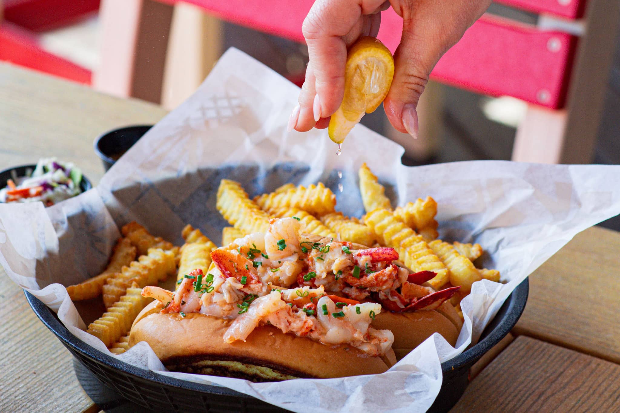 Lobster and shrimp roll topped with chives with a hand squeezing lemon over it, served with crinkle-cut fries and coleslaw